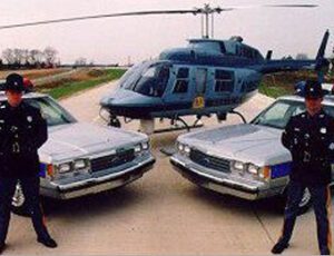 Two uniformed officers stand by their patrol cars with a helicopter in the background.